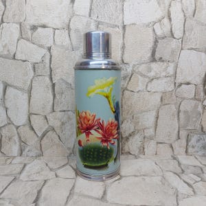 Könnte beinhalten: Eine Vintage-Thermoskanne mit hellblauem Korpus und silbernem Deckel. Die Thermoskanne hat ein Blumendesign mit gelben und roten Blüten und einem grünen Kaktus. Die Thermoskanne ist aus Metall und hält Flüssigkeiten heiß oder kalt.