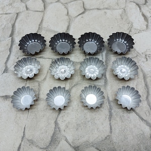 Puede incluir: Un juego de doce moldes para cupcakes de metal plateado con bordes acanalados. Los moldes están dispuestos en tres filas de cuatro sobre una superficie de piedra gris.