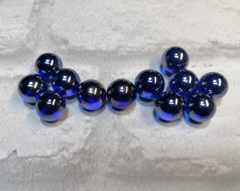 Könnte beinhalten: Eine Sammlung von irisierenden blauen Glasmurmeln. Die Murmeln haben eine reflektierende Oberfläche mit violetten und blauen Glanzlichtern. Die Murmeln sind auf einer hellgrauen Oberfläche verstreut.