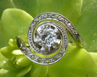 May include: A silver-toned ring featuring a central round-cut clear gemstone surrounded by a swirling metal frame with intricate cut-out details and milgrain edges, creating a spiral design that encircles the main stone.