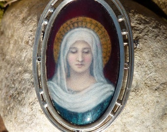 Könnte beinhalten: Ein ovales Silbermedaillon mit einem gemalten Bild einer Frau mit Heiligenschein. Die Frau trägt einen weißen Schleier und hat einen blauen und grünen Hintergrund.