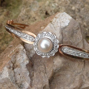 Op de afbeelding: Een goud-zilveren scharnierende armband met een centrale parel omringd door een halo van kleine, heldere edelstenen. De band van de armband heeft een rij kleine, heldere edelstenen. De armband ligt op een ruwe, lichtbruine steen.