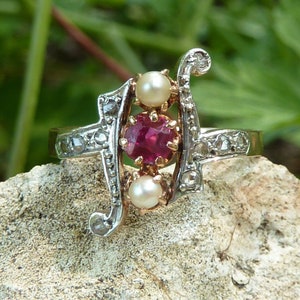 Puede incluir: Un anillo de oro y plata con un rubí y dos perlas. El anillo está engastado con pequeños diamantes y tiene un diseño delicado.
