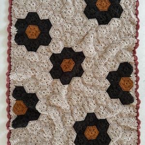 May include: A crocheted blanket with a geometric pattern of hexagons in beige, brown, and black. The blanket has a scalloped border in a burgundy color.