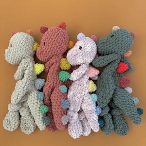 May include: Four handmade crochet dinosaur toys in shades of gray, pink, purple, and green. Each dinosaur has colorful spines and a small head. The toys are arranged on an orange background.