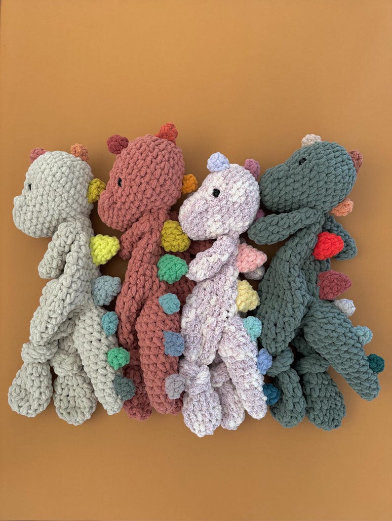 May include: Four handmade crochet dinosaur toys in shades of gray, pink, purple, and green. Each dinosaur has colorful spines and a small head. The toys are arranged on an orange background.