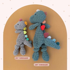May include: Two crocheted dinosaur plush toys. One is gray and measures 15 inches, the other is teal and measures 20 inches. Both have colorful spines and are labeled "SNUGGIE". The toys are displayed on a tan background with a pink dotted pattern.