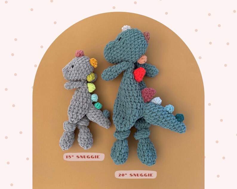 May include: Two crocheted dinosaur plush toys. One is gray and measures 15 inches, the other is teal and measures 20 inches. Both have colorful spines and are labeled "SNUGGIE". The toys are displayed on a tan background with a pink dotted pattern.