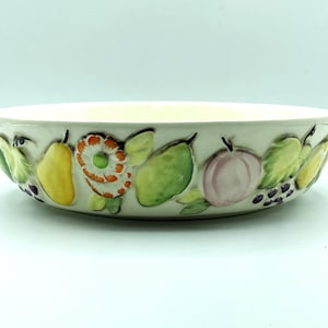 May include: A cream-colored ceramic bowl with a raised fruit and floral design around the rim. The design includes yellow pears, green leaves, a pink peach, purple grapes, and an orange and white flower. The bowl has a shallow, round shape.