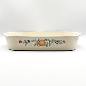 May include: A rectangular cream-colored ceramic baking dish featuring a central decorative motif of orange fruit, green leaves, and clusters of dark blue and red berries. The dish has integrated side handles and a smooth, glossy finish.