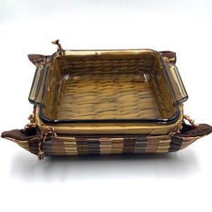 May include: A rectangular glass baking dish with a brown woven pattern, set inside a matching woven basket-style holder. The holder has brown fabric accents and decorative rope ties. The dish is designed for baking and serving.