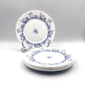 May include: White ceramic plates with a blue floral design. The plates have a delicate blue floral border and a small floral accent in the center. One plate is displayed on a black metal stand, and a stack of plates is visible.