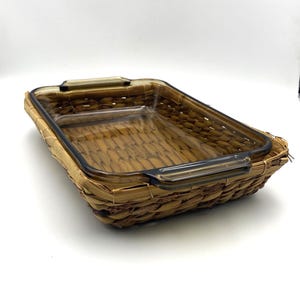 May include: A rectangular glass baking dish with dark brown handles sits inside a woven basket. The basket is made of natural-colored woven material. The dish is empty and ready for use. The background is white.