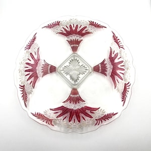 May include: A clear glass serving platter with a decorative floral design in red and white. The platter has a scalloped edge and a central square compartment. The floral design radiates from the center, creating a symmetrical pattern.