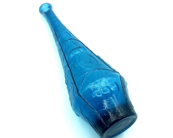 May include: A tall, slender, cobalt blue glass bottle featuring a narrow neck and a wider, rounded base. The exterior surface of the bottle is decorated with a repeating embossed floral pattern that covers the body from the base to the neck.