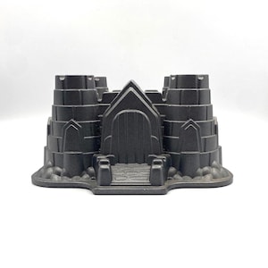 May include: A dark gray castle-shaped cake pan with a detailed design. The pan features a central arched doorway, two towers, and a tiered structure. The pan is made of a sturdy material and is designed for baking decorative cakes.
