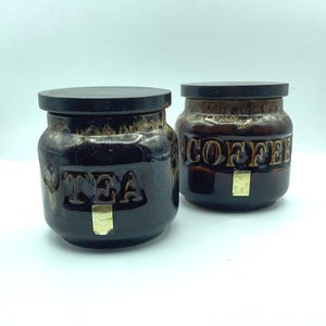 May include: Two ceramic tea and coffee canisters. They are dark brown with black lids and gold accents. One canister has the word "TEA" embossed, the other "COFFEE".
