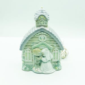 May include: A ceramic wedding chapel figurine in pastel colors. The chapel has a gray roof and green trim, with a bride and groom figurine in front. The figurine is decorated with floral accents and has a delicate, romantic aesthetic.