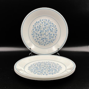 May include: A set of white ceramic plates with a blue floral pattern. Each plate has a blue border and a central design of small blue flowers. The plates are stacked and displayed on a black surface.