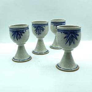 May include: Four handmade ceramic goblets. Each goblet has a speckled, off-white body with a blue floral design and a blue band around the rim. The base is a light brown color. The goblets are displayed on a white surface.