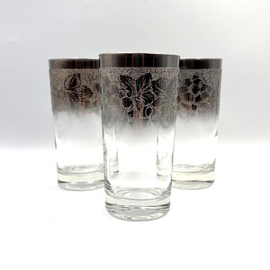 May include: Three clear glass tumblers featuring a metallic silver band around the top rim. Each glass is decorated with a frosted silver etched pattern of grape clusters and leaves encircling the upper portion of the cylindrical vessel.