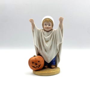 May include: A ceramic figurine of a child dressed as a ghost, with arms raised. The figure wears a white sheet and a matching hood. A carved orange pumpkin sits at the child's feet. The figurine is on a round, tan base.