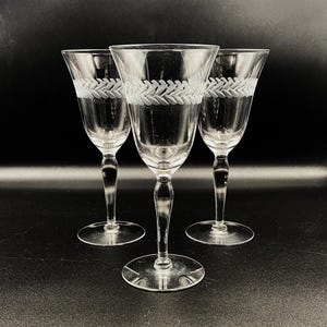 May include: Three clear wine glasses with a decorative etched leaf pattern around the bowl. The glasses have a long stem and a round base. The background is black.
