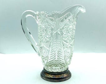 May include: A clear glass pitcher with a starburst pattern and a silver-colored base. The pitcher has a handle and a spout for pouring. The design is intricate and detailed, suitable for serving beverages.