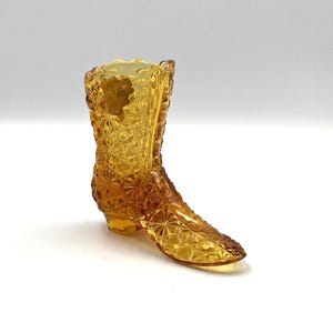 May include: A decorative amber-colored glass boot with a textured, starburst pattern. The boot has a pointed toe and a small heel. The glass is transparent, allowing light to pass through, and is likely a collectible or decorative item.