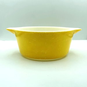 May include: A round, bright yellow casserole dish with a smooth, glossy finish and two integrated side handles. The interior of the bowl is white, contrasting with the vibrant yellow exterior. The dish features a wide, open top design.