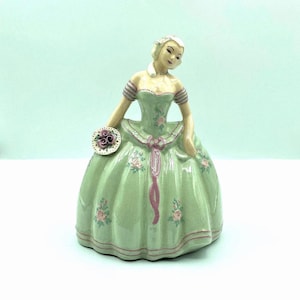 May include: A vintage porcelain figurine of a woman in a long, light green dress adorned with pink roses and a matching ribbon. The figurine holds a small, round plate with a floral design. The woman has blonde hair and is wearing a matching headpiece.