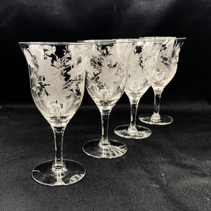 May include: Four clear glass wine glasses with etched floral designs. The glasses are arranged in a row on a black surface.