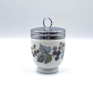 May include: A white ceramic egg cup with a silver-colored lid and handle. The cup is decorated with a floral pattern featuring green leaves and purple berries. The egg cup is designed to hold a single egg.