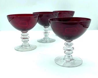May include: Four ruby red stemmed glasses with a rounded bowl and a clear, decorative stem. The glasses are arranged on a white surface, showcasing their elegant design. Perfect for serving cocktails or champagne.