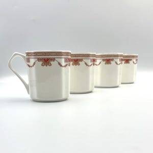 May include: A set of four white ceramic mugs arranged in a row. Each mug features a cylindrical body with a smooth finish and a decorative border near the rim, consisting of thin brown lines, small red floral accents, and repeating geometric patterns.