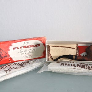 Pode incluir: Uma caixa vermelha e branca com o texto "The Everyman London Pipe" e uma imagem de um homem fumando um cachimbo. A caixa contém um cachimbo de madeira marrom com uma haste preta e uma bolsa de tecido branco. Também estão incluídos dois pacotes de limpadores de cachimbo brancos.