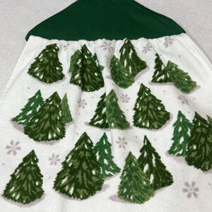 May include: White hanging kitchen towel with a green top and a green button. The towel features a pattern of green Christmas trees and small gray snowflakes. The towel is made of a soft, absorbent material.