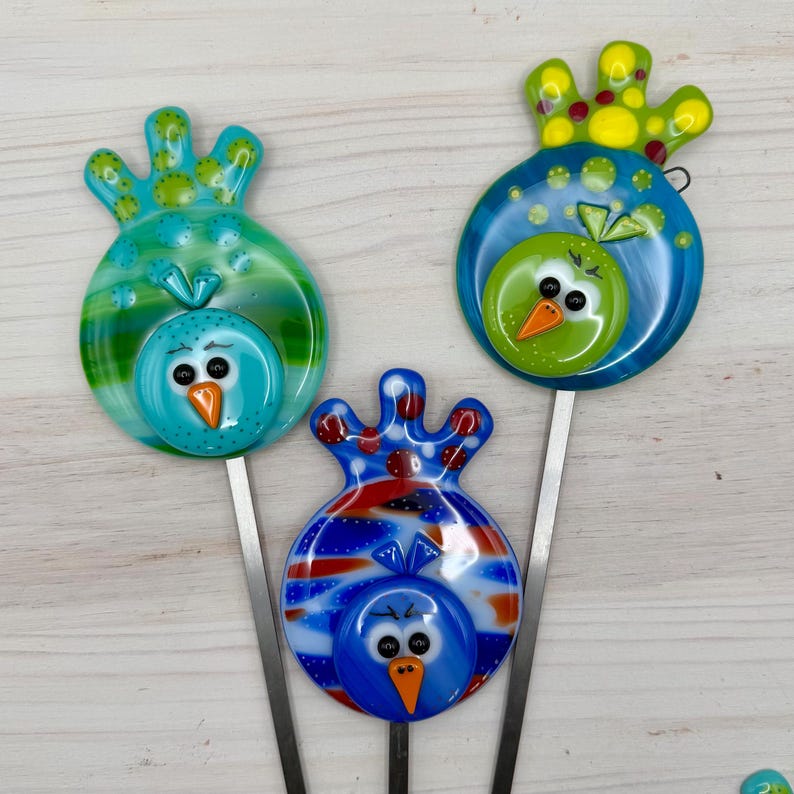 May include: Three colorful glass bird garden stakes. The birds have round bodies and decorative headpieces. The birds are blue, green, and red with orange beaks and black eyes. The stakes are made of metal.
