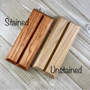 Op de afbeelding: Twee houten displaystandaards, &eacute;&eacute;n gebeitst in een warme bruine kleur en de andere ongebeitst, natuurlijk hout. Elke standaard heeft een groef voor het vasthouden van items. Het woord "Stained" staat in zwart script boven de gebeitste standaard, en "Unstained" staat eronder.