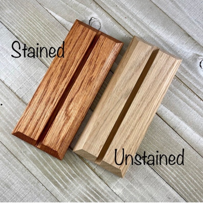 Op de afbeelding: Twee houten displaystandaards, &eacute;&eacute;n gebeitst in een warme bruine kleur en de andere ongebeitst, natuurlijk hout. Elke standaard heeft een groef voor het vasthouden van items. Het woord "Stained" staat in zwart script boven de gebeitste standaard, en "Unstained" staat eronder.