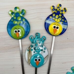 May include: Three colorful glass bird garden stakes. The birds have round bodies and decorative headpieces. One bird is green with a blue headpiece, another is yellow with a blue headpiece, and the third is turquoise with a white and brown headpiece.