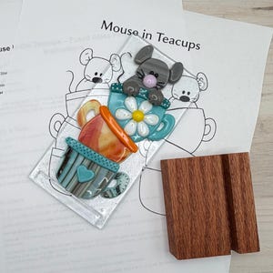 Peut inclure: Pièce d'art en verre fusionné intitulée "Mouse in Teacups" représentant une souris grise sur une tasse bleue avec une marguerite blanche. En dessous, deux autres tasses en orange et sarcelle. Un support en bois est à côté de l'œuvre. Instructions en arrière-plan.