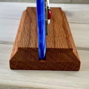 Op de afbeelding: Een handgemaakte houten telefoonstandaard met een sleuf om een telefoon in te plaatsen. De standaard is gemaakt van roodbruin hout en heeft een decoratief glazen element met blauwe, witte en rode accenten. Het glazen element heeft een lieveheersbeestje.