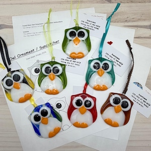 Peut inclure: Plusieurs ornements en verre fusionné en forme de hibou, de couleurs variées : vert, bleu, rouge et marron. Chaque ornement présente un ventre blanc, des pieds orange et de grands yeux noirs. Ils sont suspendus par des rubans et accompagnés de cartes imprimées.