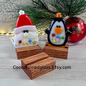 May include: Two stained glass Christmas ornaments, a Santa Claus and a penguin, on wooden stands. The ornaments are decorated with colorful lights and beads. The stands are made of dark wood with a natural grain.