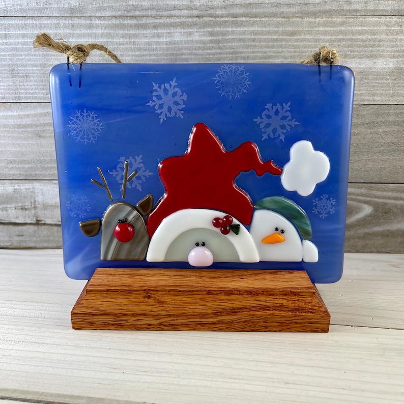 K&ouml;nnte beinhalten: Blaue Weihnachtsdekoration aus Glas mit Rentier, Wichtel und Schneemann. Die Figuren sind rot, wei&szlig; und gr&uuml;n, mit Schneeflocken-Akzenten. Auf einem Holzst&auml;nder montiert, mit einer Juteschnur zum Aufh&auml;ngen.