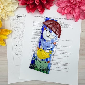 May include: A fused glass panel featuring four colorful birds stacked vertically. The top bird is under a red umbrella. The panel has a blue background with white stripes. Instructions and a sketch are in the background.