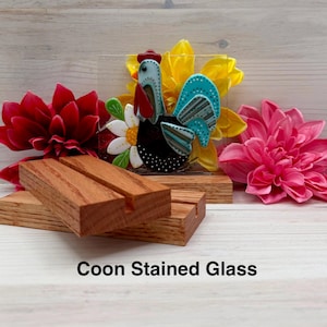 May include: A colorful stained glass rooster with a red comb and turquoise wings, set against a yellow flower and a white daisy. The rooster is displayed on a wooden base, with two additional wooden blocks in the foreground. The text "Coon Stained Glass" is at the bottom.