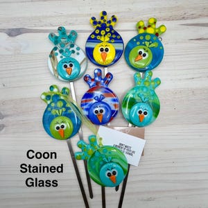May include: A collection of colorful, stained glass bird garden stakes. Each bird features a unique design with vibrant blues, greens, yellows, and oranges. The text "Coon Stained Glass" is visible. The stakes are on metal rods.