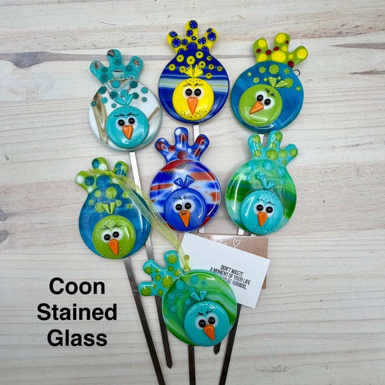 May include: A collection of colorful, stained glass bird garden stakes. Each bird features a unique design with vibrant blues, greens, yellows, and oranges. The text "Coon Stained Glass" is visible. The stakes are on metal rods.